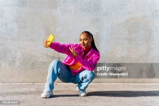 smiling young dancer in squatting position taking selfie through mobile phone in front of wall - coolpad stockfoto's en -beelden