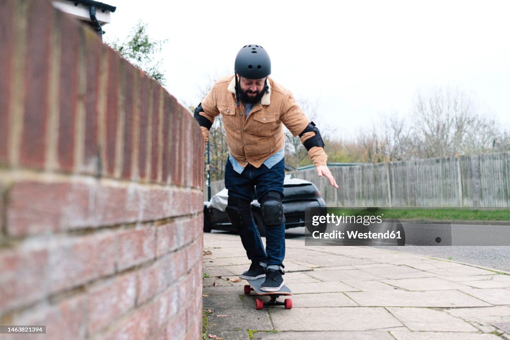 Mature man learning skateboarding taking support of wall on footpath