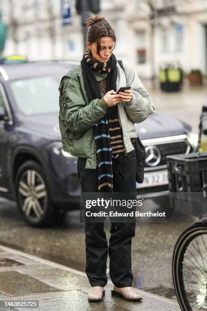 Guest wears a black with beige embroidered pattern wool long scarf, a khaki nylon oversized bomber coat, a brown wool pullover, black suit pants,...