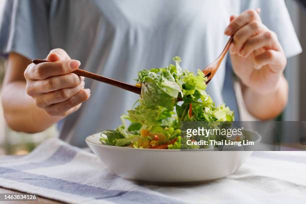 young asian woman mixing ingredients in her healthy fresh vegan salad - saladekom stockfoto's en -beelden