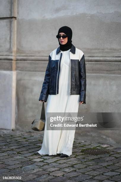 Guest wears a black wool balaclava, black sunglasses, a white pleated long coat, a black and white bicolored shiny leather zipper jacket, a brown...