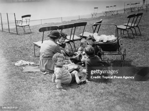 Nanny or childminder with a group of five young multiracial children sitting on grass while having a picnic, there is a baby pram parked beside them,...