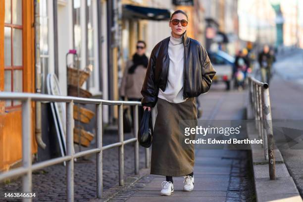 Guest wears black and orange sunglasses, a white ribbed wool turtleneck pullover, a black shiny leather bomber coat, a dark gray long skirt, black...