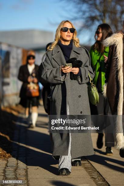 Guest wears black sunglasses, a navy blue ribbed wool turtleneck pullover, a gray long wool coat, black gloves, a green shiny leather Saddle handbag...