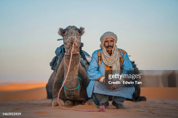 moroccan camel driver squatting in sahara desert with camel looking at camera - morocco stock pictures, royalty-free photos & images