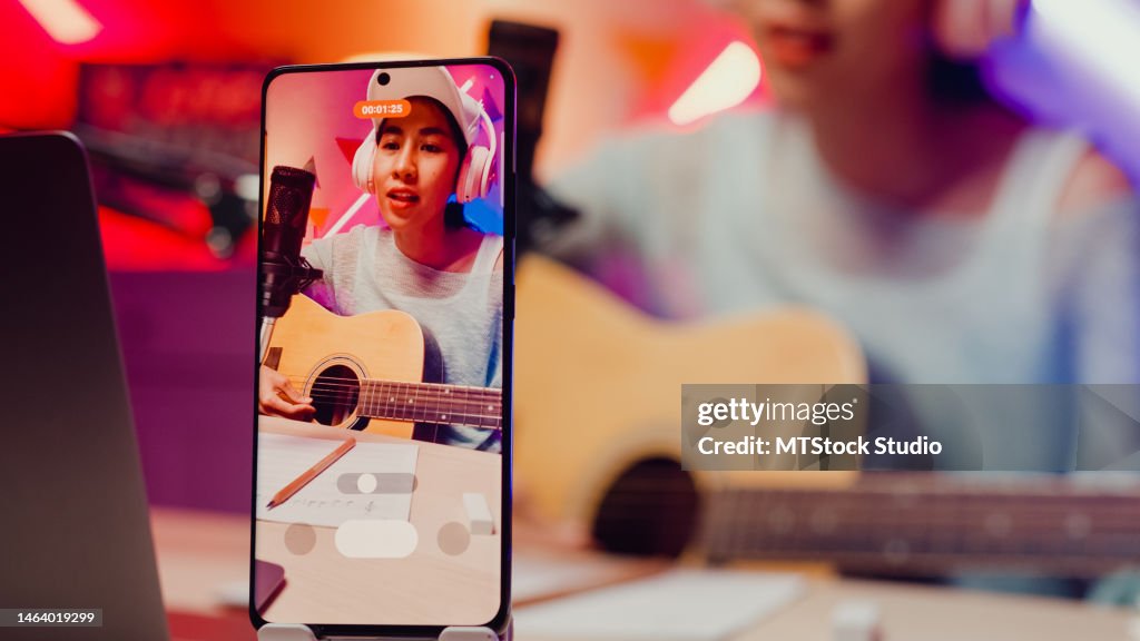 Close-up Happy Asian musician girl influencer wear headphone play guitar record phone podcast online live stream sing song with follower audient at studio.