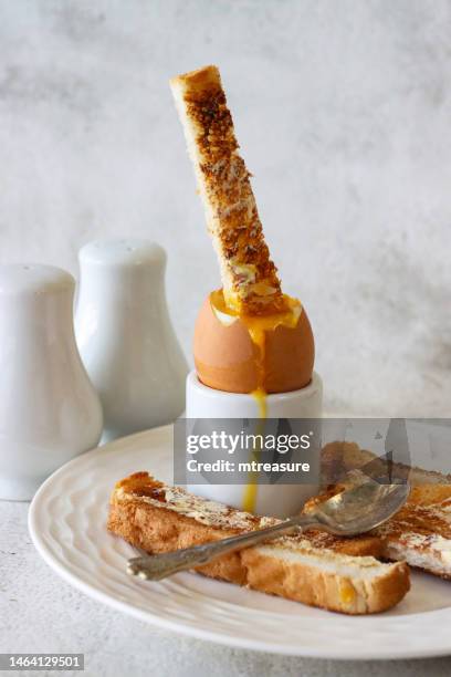 image of white buttered toasted bread slice being dipped into runny yellow yolk of soft boiled egg, white eggcup, metal teaspoon, salt and pepper shaker cruet set, marble effect background, focus on foreground - hard boiled eggs stock pictures, royalty-free photos & images