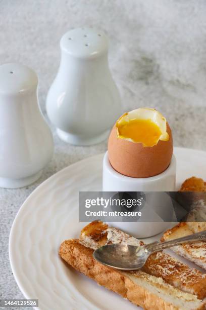 close-up image of soft boiled egg with soldiers, runny yolk, white bread toast slices with butter, white eggcup, metal teaspoon, salt and pepper shaker cruet set, marble effect background, focus on foreground - cruet stock pictures, royalty-free photos & images