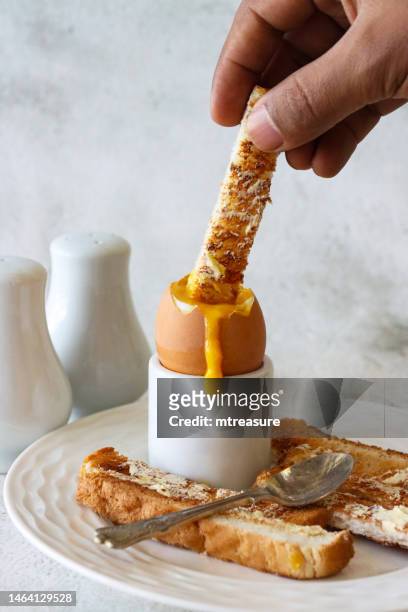 image of white buttered toasted bread slice being dipped into runny yellow yolk of soft boiled egg by unrecognisable person, white eggcup, metal teaspoon, salt and pepper shaker cruet set, marble effect background, focus on foreground - bread butter stock pictures, royalty-free photos & images