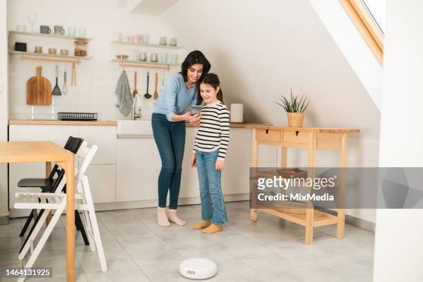 woman and her little girl using smartphone and turning on their smart vacuum cleaner - modern family media call stockfoto's en -beelden
