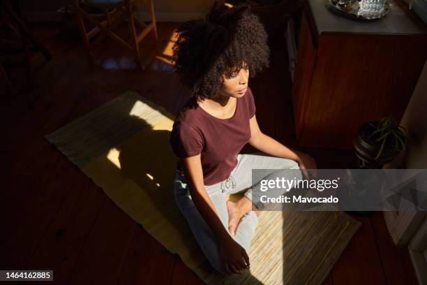 young african woman meditating by a sunny window at home - gambe incrociate foto e immagini stock