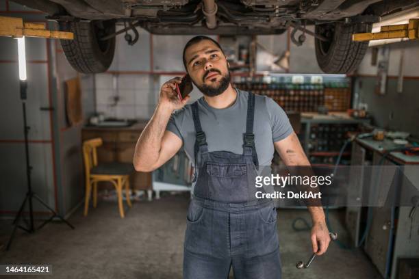 car mechanic talking on mobile phone while repairing broken vehicle in auto repair shop - repairman phone stock pictures, royalty-free photos & images