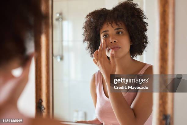 young african woman putting cream on her face in a bathroom mirror - trattamento per la pelle foto e immagini stock
