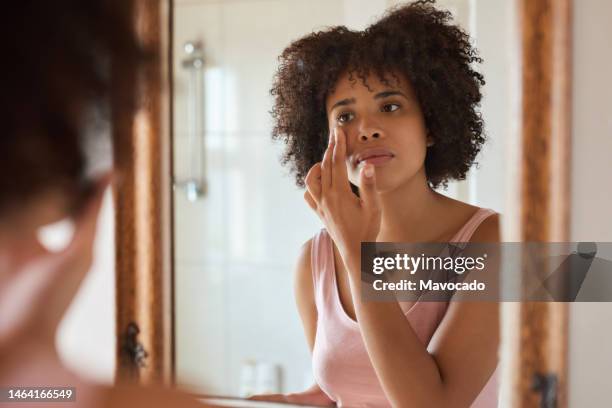 young african woman putting cream on her face in a bathroom mirror - applying stockfoto's en -beelden