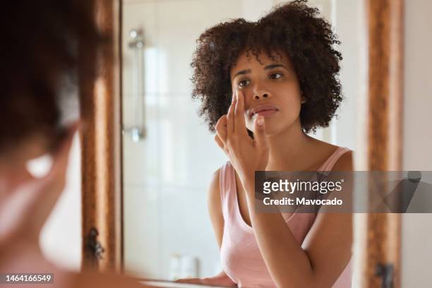 young african woman putting cream on her face in a bathroom mirror - applying imagens e fotografias de stock