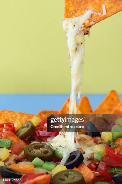 full frame image of unrecognisable person holding nacho, plate of loaded nachos covered in tomato salsa, melted mozzarella cheese, tomatoes, black olives, bell peppers, jalapeno peppers, red chillies, split yellow-blue background, focus on foreground - black olive stock pictures, royalty-free photos & images