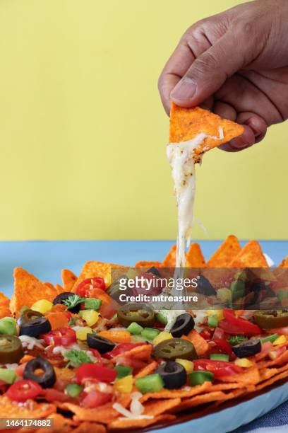 close-up image of unrecognisable person holding nacho, plate of loaded nachos covered in tomato salsa, melted mozzarella cheese, tomatoes, black olives, bell peppers, jalapeno peppers, red chillies, split yellow-blue background, focus on foreground - black olive stock pictures, royalty-free photos & images