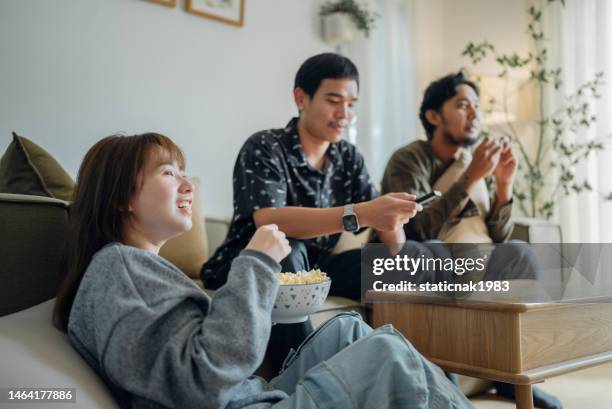 young asian relaxing eating corn snacks while watching movie or tv on sofa in living room at home. - asian watching tv stock pictures, royalty-free photos & images