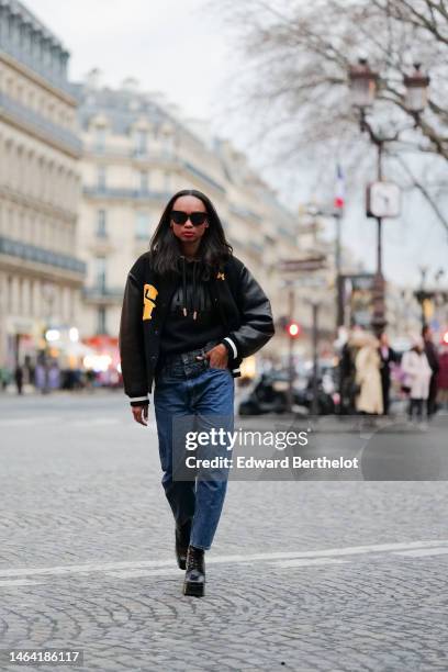 Emilie Joseph wears black sunglasses, a black hoodie sweater from Ami, a black felt and leather sleeves with embroidered yellow yoke oversized teddy...