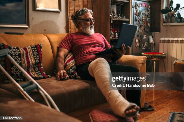 mature man with broken leg in plaster cast on sofa - broken laptop stockfoto's en -beelden