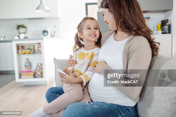 pregnant mother and her little daughter enjoying at home and using a smart phone - little kids belly imagens e fotografias de stock