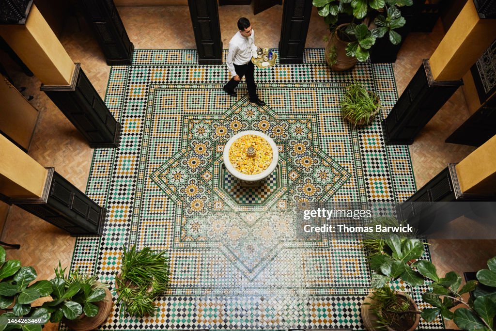 Wide shot of waiter carrying tea service through courtyard in hotel