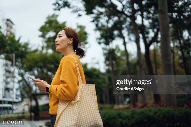 young asian woman carrying a reusable shopping bag, using smartphone while walking in the city against trees outdoors. responsible shopping, zero waste, eco-friendly concept. sustainable living lifestyle - tote bag stock pictures, royalty-free photos & images