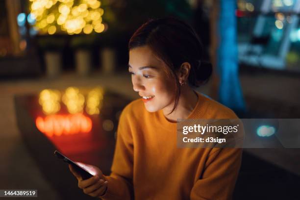 beautiful smiling young asian woman sitting in a pub and relaxing, surfing on the net and checking social media on smartphone. spending a night out and enjoying city night life. lifestyle and technology - bank nightclub stock pictures, royalty-free photos & images