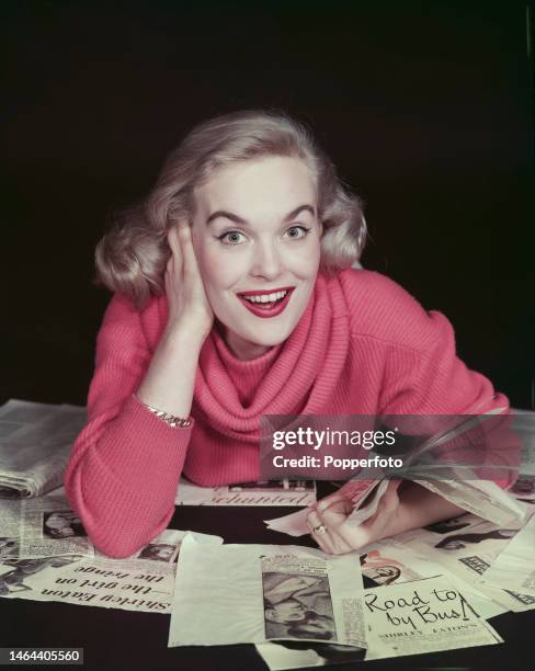 English actress Shirley Eaton posed wearing a pink ribbed sweater with a cowl neck with an assortment of press cuttings in London on 16th April 1957.