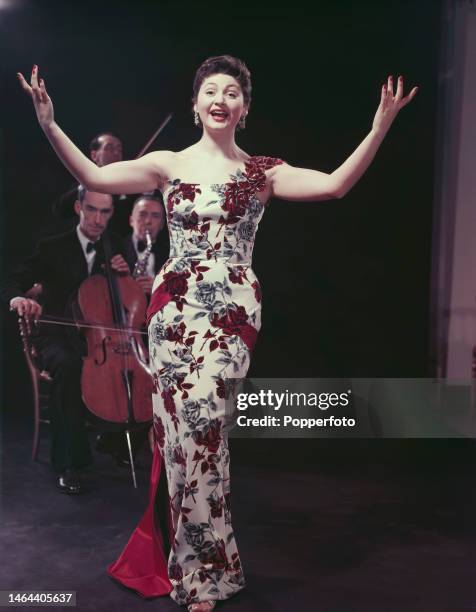 Posed studio portrait of English singer and operatic soprano Adele Leigh wearing a full length floral dress with rose motif, and a pink satin...