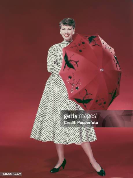 Posed studio portrait of a woman wearing a mid length, full skirted white dress featuring a black circle pattern, she holds a red umbrella with black...