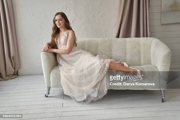 a beautiful young bride in a wedding dress is sitting on the sofa in a room with a beautiful interior with curtains dividing the room. a woman in a full-length long dress and shoes. a lifestyle with a copy space. - fabulous full lengths foto e immagini stock