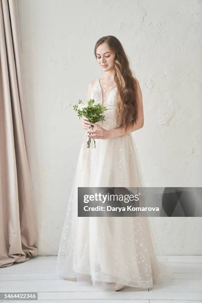 a beautiful young bride in a wedding dress and flowers standing in a room with a beautiful interior with curtains. a woman in a full-length long dress. a lifestyle with a copy space. - fabulous full lengths foto e immagini stock