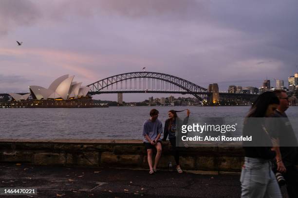 Woman playing with her hair talks to a man sitting at Mrs Macquaries Point with a view of Opera House and Harbour Bridge in the background on...