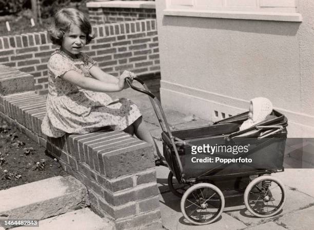 British Royal Princess Elizabeth of York, wearing a short sleeve dress, sits on a brick wall, with her hands on the handlebar of a pram, in which is...