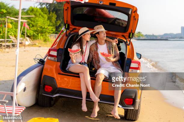 relaxing young couple holding a watermelon sitting behind of a car at the sea. - holiday healthy eating stock pictures, royalty-free photos & images