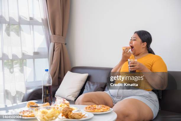 plus size woman sitting on sofa and eating junk food. - fat woman stock-fotos und bilder