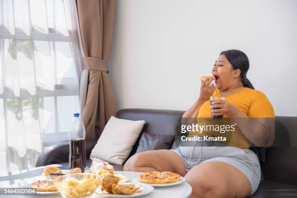 plus size woman sitting on sofa and eating junk food. - fat people eating donuts foto e immagini stock