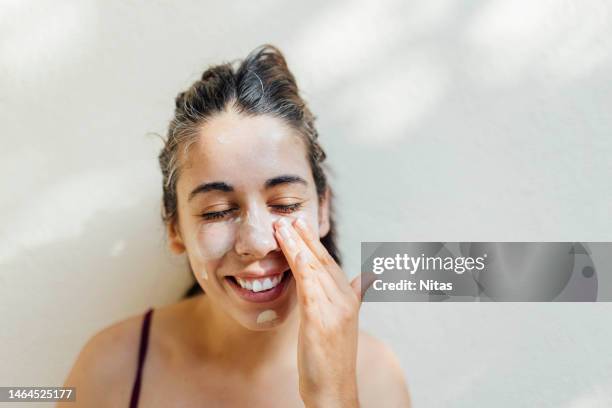 a close-up portrait of a young, happy hispanic woman applying sunscreen to her cheeks and forehead - frau gesicht beauty treatment stock-fotos und bilder