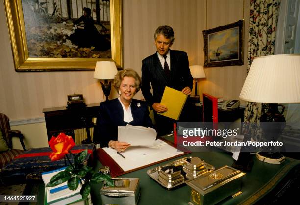 Conservative Party politician and Prime Minister of the United Kingdom Margaret Thatcher seated signing documents at a desk with Private Secretary...
