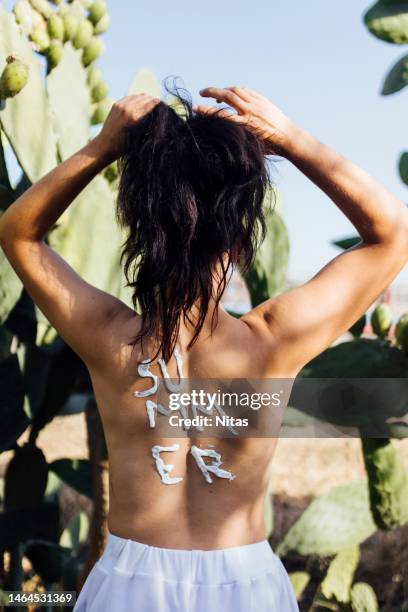 rear view of young brunette millennial woman on her back with sun tan lotion on her back for protect her skin in front of a cactus. skincare and summer - summer hair care stock pictures, royalty-free photos & images