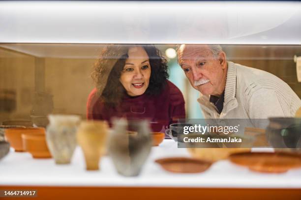 a senior caucasian couple looking at a glass display cabinet with pottery vessels - couple art gallery stock pictures, royalty-free photos & images