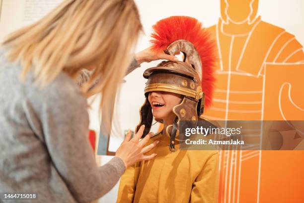 blonde caucasian mother putting roman galea on her happy young son's head while they are visiting a museum - classical imagens e fotografias de stock