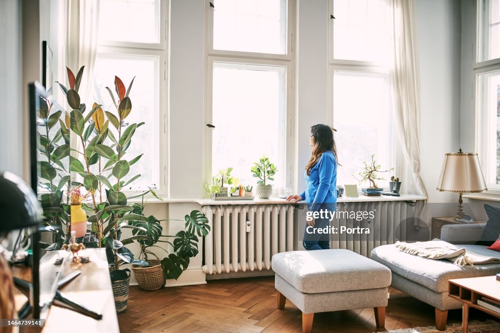 Mid adult woman standing by radiator and looking outside the window at home