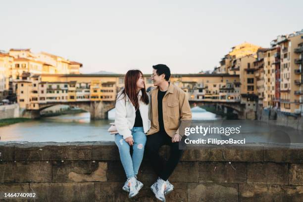 ein asiatisches paar sitzt auf der brücke santa trinita mit der ponte vecchio im hintergrund - florenz, italien - vecchio stock-fotos und bilder