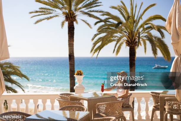 young blonde woman sitting in the beach restaurant enjoying a red cocktail with the turquoise sea and palm trees and a yacht in the background - spanje stockfoto's en -beelden