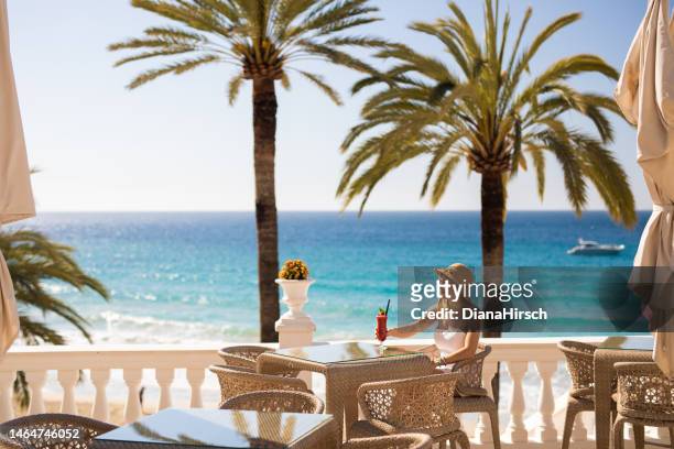 young blonde woman sitting in the beach restaurant enjoying a red cocktail with the turquoise sea and palm trees and a yacht in the background - maiorca 個照片及圖片檔