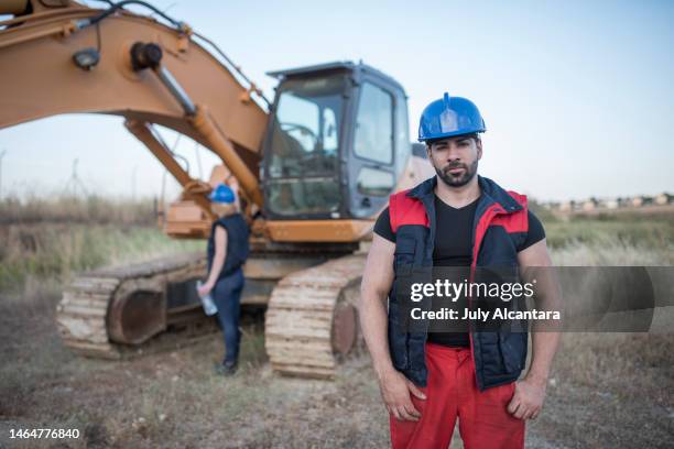 trabajador de la construcción fuerte cerca de la retroexcavadora mirando a la cámara, mujer en el fondo - pregnant july 2017 fotografías e imágenes de stock