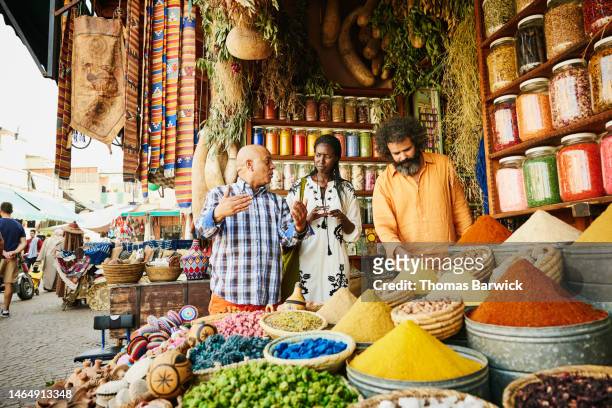 wide shot couple talking to spice shop owner in the souks of marrakech - marrakech spice stockfoto's en -beelden