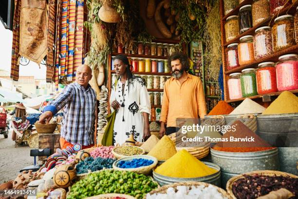 wide shot couple talking to spice shop owner in the souks of marrakech - morocco tourist stock pictures, royalty-free photos & images